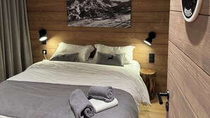 A neatly made bed with a grey blanket and two pillows, a wooden wall with a number sign, a mounted picture of mountains, and two wall-mounted lamps.