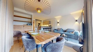 A modern living room with a wooden dining table, grey upholstered chairs, a grey sofa, and a kitchen in the background.
