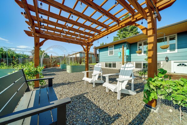 A wooden pergola with hanging lights, a bench, and white Adirondack chairs in a garden area with a house in the background. - 3 Mi to Dtwn: Family Home w/ Garden & EV Charger (Yakima)