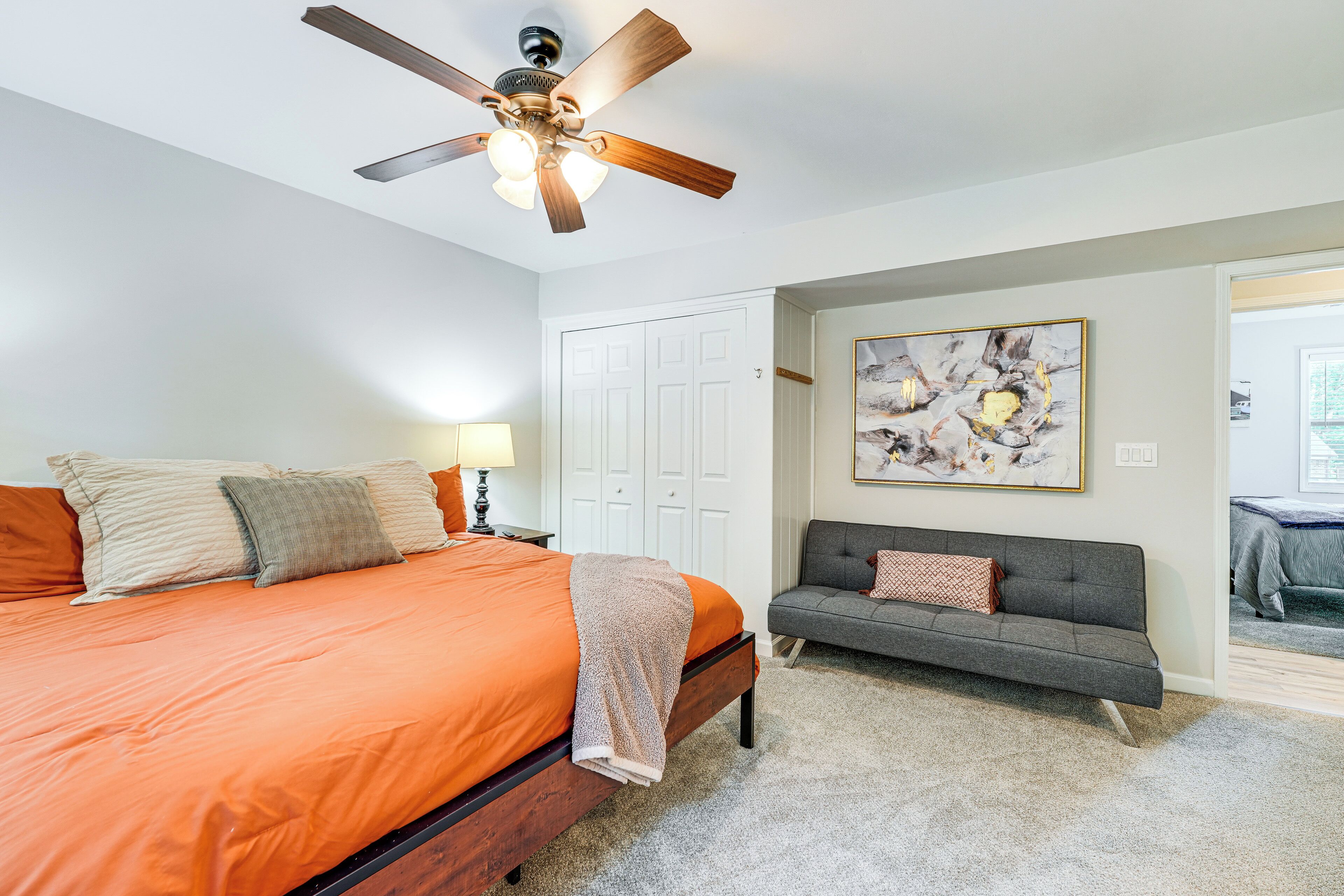 A bedroom with a bed, a gray sofa, a ceiling fan, a white door, and a framed abstract painting on the wall.