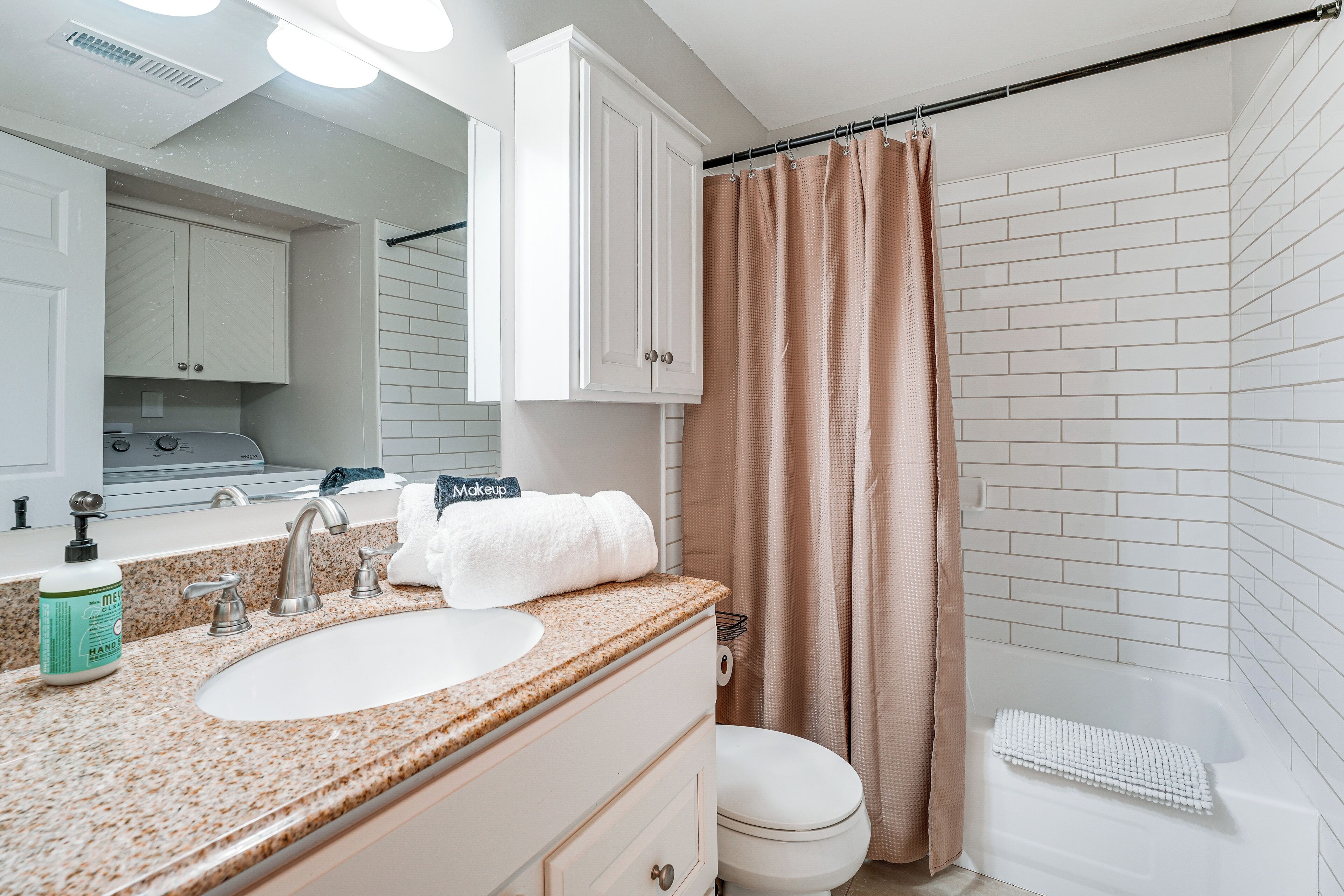 Ein Badezimmer mit weißem Waschbecken, Waschtisch und Toilette. Ein Duschvorhang, ein Handtuchhalter und eine Handseifenflasche befinden sich auf der Ablagefläche.