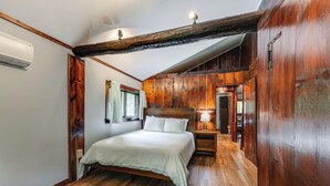 Une chambre avec un plafond en bois, un lit recouvert de linge de lit blanc, une table de chevet avec une lampe et une fenêtre avec des rideaux.