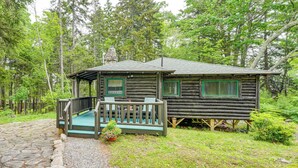 Une cabane rustique en rondins de bois, dotée d’un porche et de balustrades vertes, entourée d’une forêt.