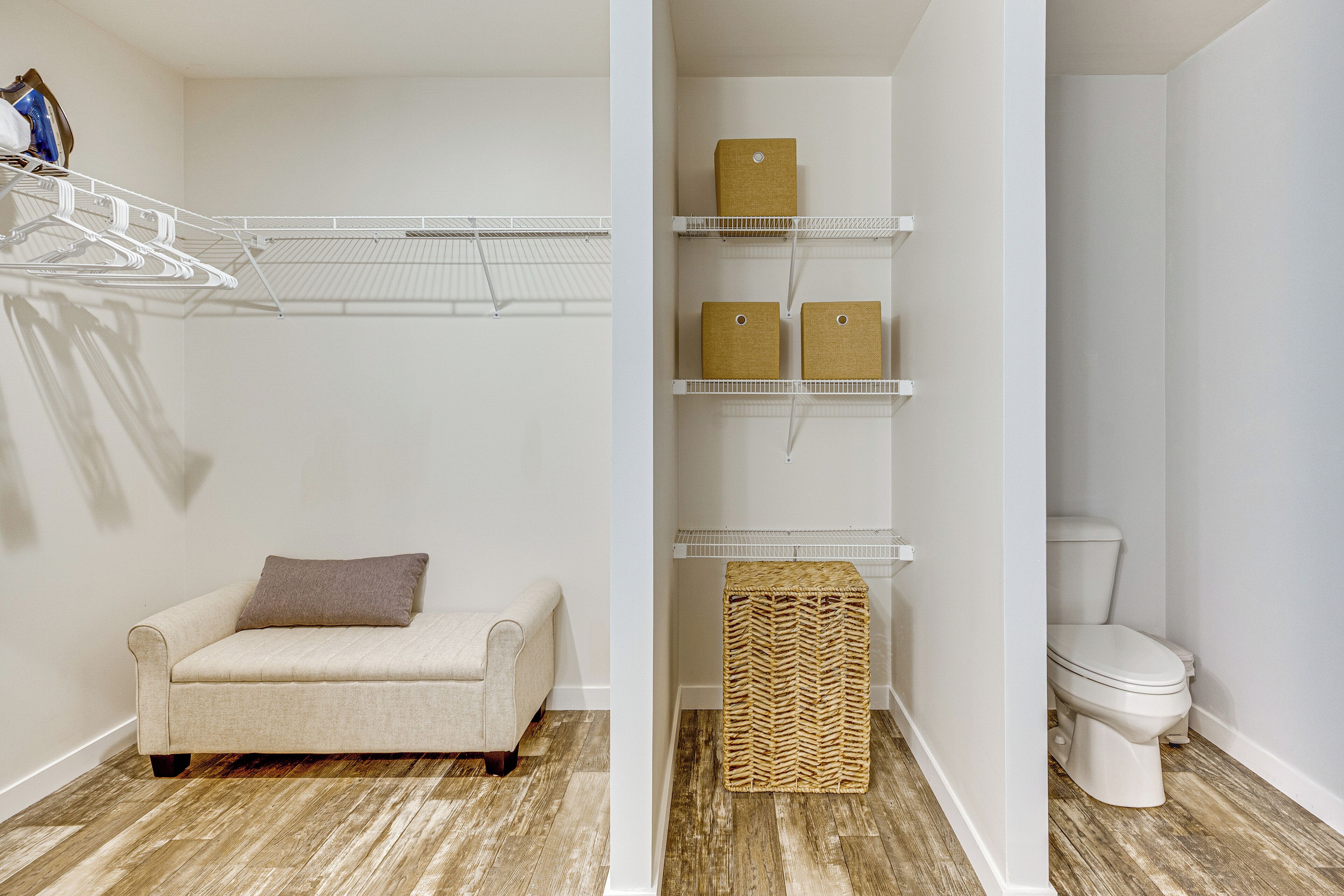 Una habitación con un sofá color beige, una pared blanca, piso de madera, inodoro blanco y una estantería metálica con cajas.
