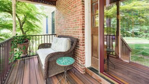 A wooden deck with a wicker sofa and a small table, a brick wall, and a large window overlooking a green landscape. - Peaceful Country Retreat: Private & Convenient (Frenchtown)
