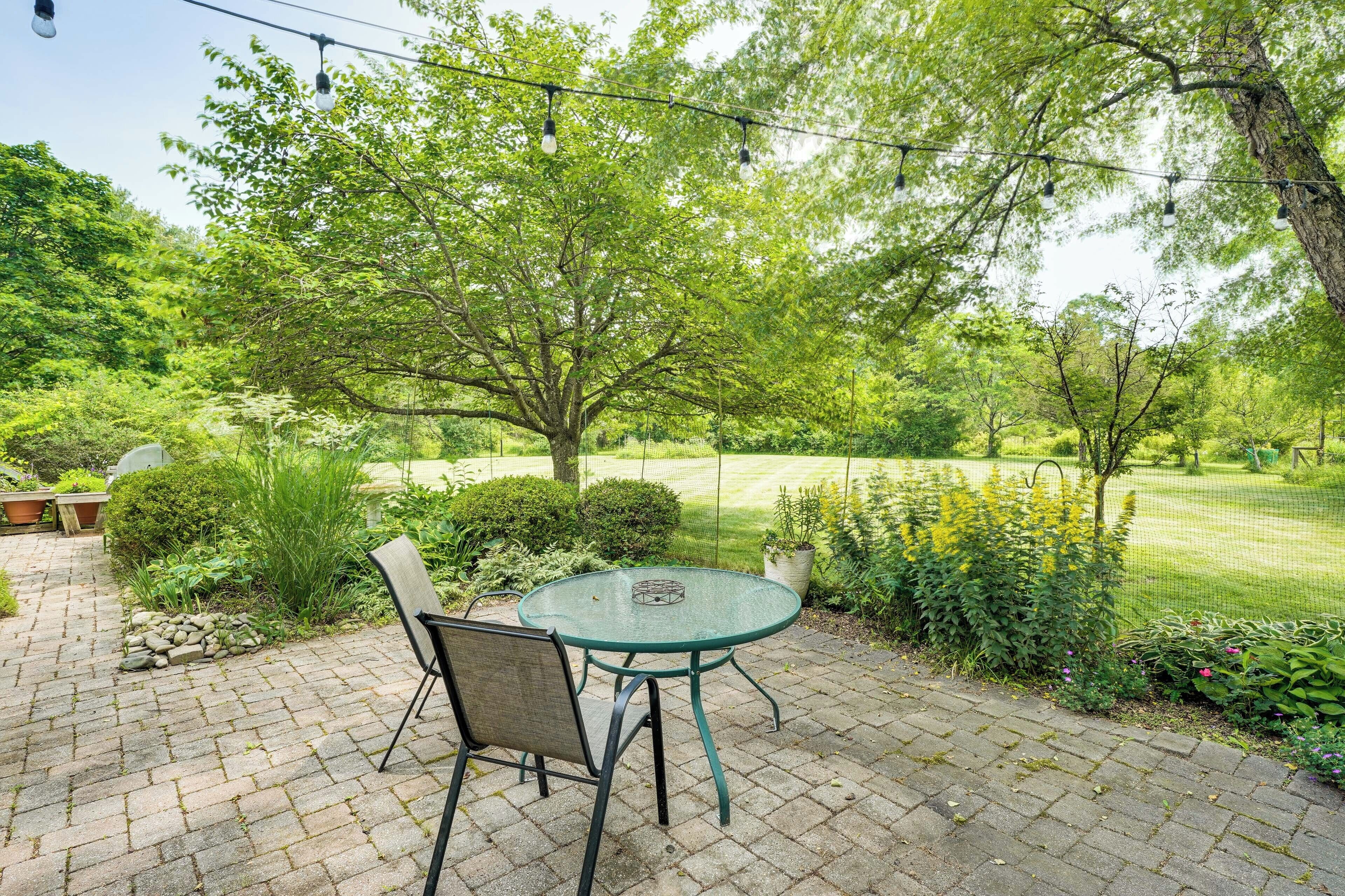 An outdoor patio with a round table and chairs, surrounded by lush greenery and a tree.