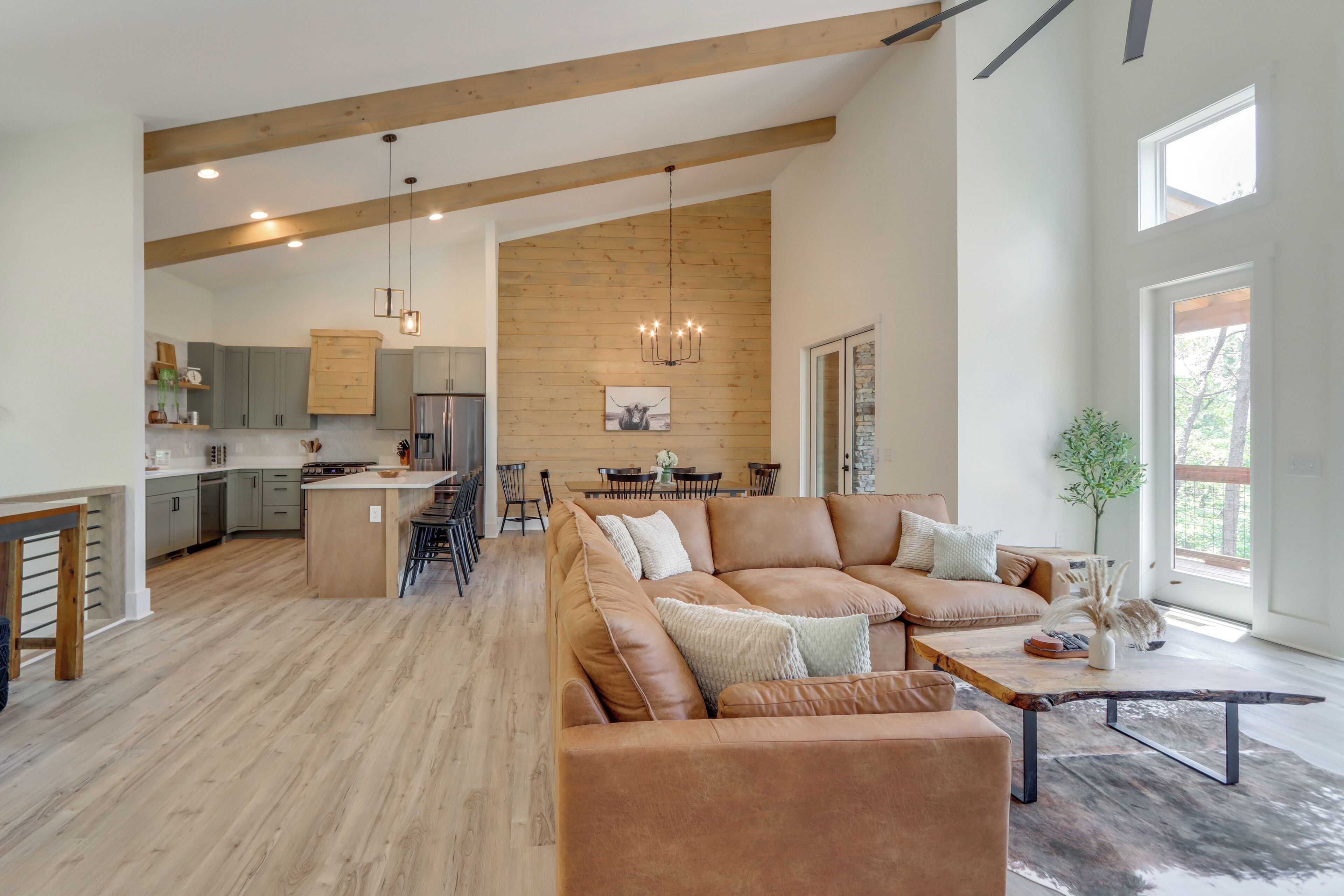 A spacious living area with a large brown leather sofa, a wooden coffee table, and a dining table with chairs. The kitchen is visible in the background with wooden cabinets and a hanging light fixture.