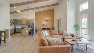 A spacious living area with a large brown leather sofa, a wooden coffee table, and a dining table with chairs. The kitchen is visible in the background with wooden cabinets and a hanging light fixture.