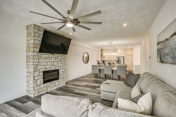 A modern living room with a stone fireplace, a flat-screen TV, a ceiling fan, and a dining area in the background. - Walk to Mississippi River: Grafton Townhome! (Grafton)