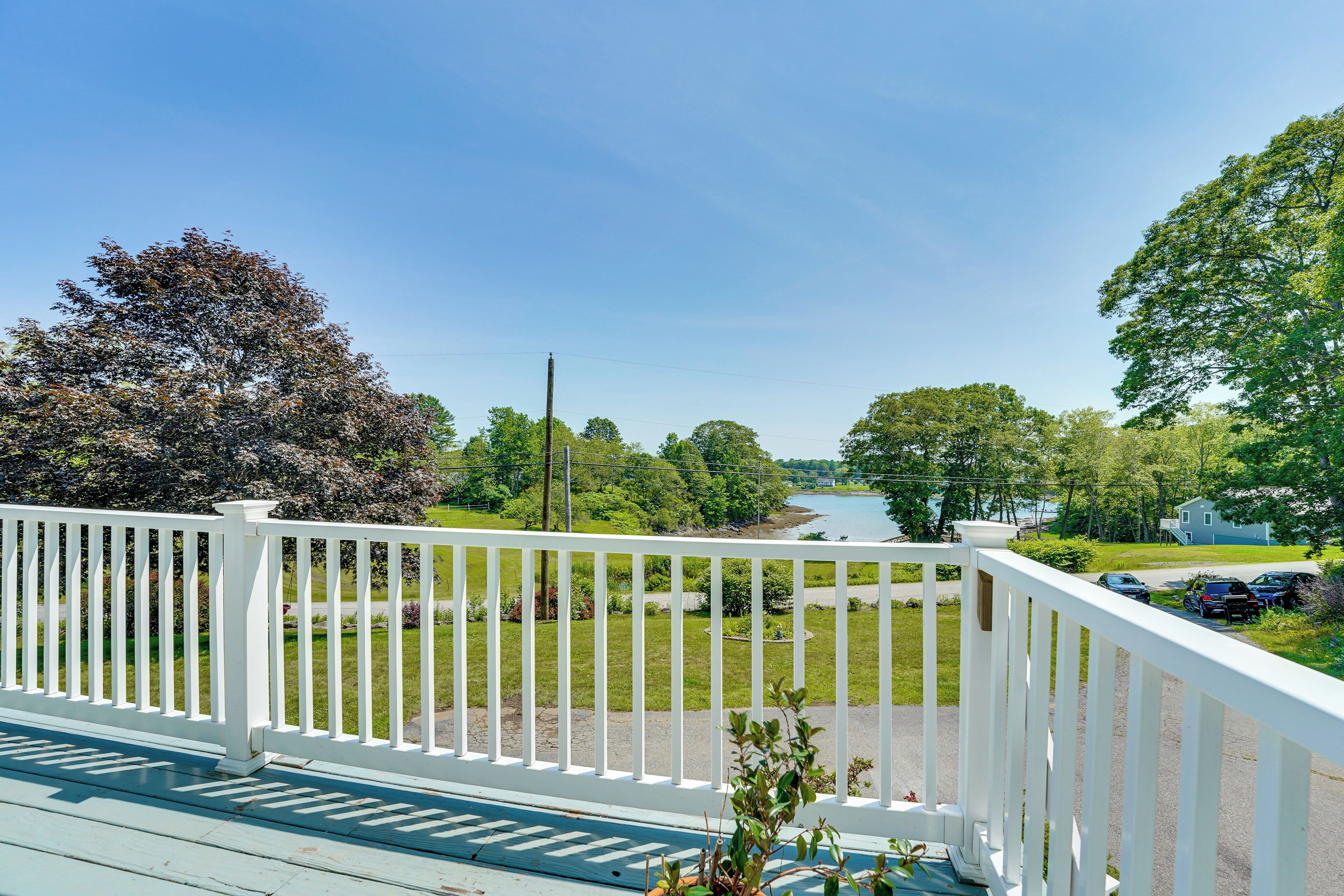 A view from a balcony with a white railing, overlooking a river, greenery, and a house with parked cars.