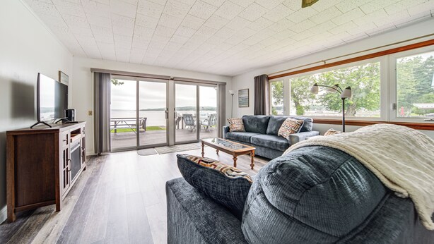Ett rymligt vardagsrum med en stor soffa, ett soffbord och en tv som står på ett träskåp. Det finns en skjutdörr i glas som leder ut till en uteplats med utsikt över vattnet.