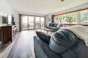 Amplia sala de estar con un sofá grande, una mesa de centro y un televisor sobre un mueble de madera. Posee un ventanal corredero que da a una terraza exterior con vista al agua.