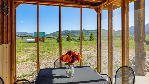 Eine Holzkonstruktion mit Tisch und Stühlen, einer Vase mit Blumen und Blick auf ein grünes Feld und Berge.