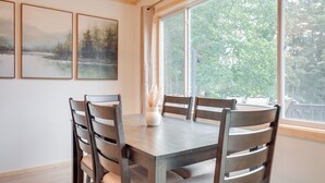 A dining area with a wooden table and chairs, framed artwork on the wall, and a large window with a view of trees. - Lake Escape on Post Lake With Private Dock (Elcho)