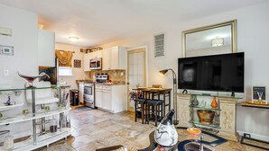 A modern kitchen with white cabinets, a dining area with a table and chairs, a TV mounted on the wall, and a glass coffee table with decorative items. - 8 Mi to Long Beach: Cozy Apt w/ Fenced Yard! (Baldwin)