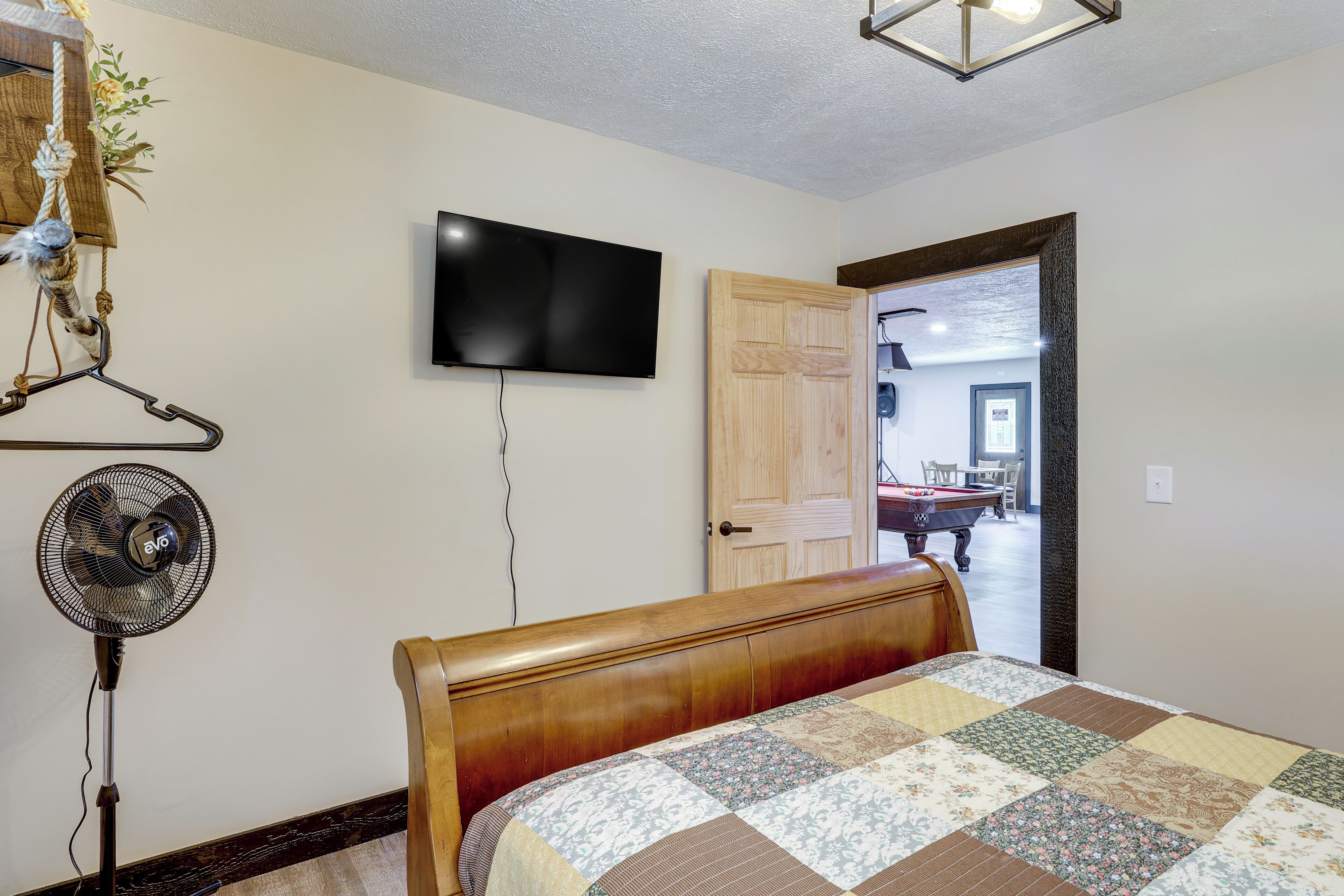 A bedroom with a wooden bed, a quilt, a wall-mounted TV, a fan, and a door leading to another room.