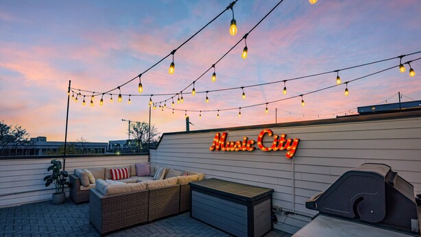 An outdoor patio area with string lights, a sofa, a grill, and a neon sign that reads "Music City". - Ultimate Rooftop Double Games BBQ Skyline Views (Nashville)