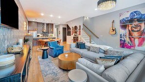 A modern living room with a large sofa, coffee table, and blue chairs. There is a kitchen area in the background with a dining table and chairs. - Ultimate Rooftop Double Games BBQ Skyline Views (Nashville)