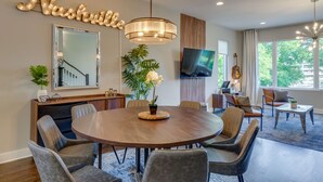 A modern dining room with a round wooden table, grey upholstered chairs, a large mirror, a potted plant, and a television on the wall. - Ultimate Rooftop Double Games BBQ Skyline Views (Nashville)
