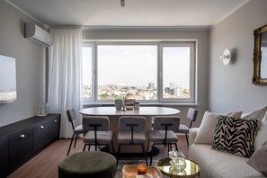 A modern living room with a dining area, large windows with a city view, a sofa with patterned cushions, and a round table with chairs. - Views of all the Palaces 2BR Stunner (Bucharest)