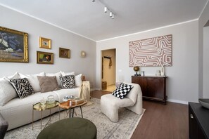 A modern living room with a white sofa, a small round table, and a decorative chair. There are framed pictures on the wall and a large abstract painting. - Views of all the Palaces 2BR Stunner (Bucharest)