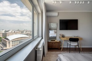 A modern hotel room with a large window, a bed, a desk with a chair, a flat-screen TV, and a radiator. - Views of all the Palaces 2BR Stunner (Bucharest)