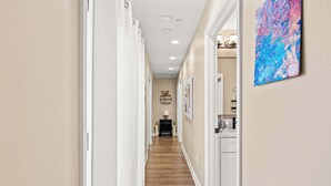 A hallway with a wooden floor, white walls, and a ceiling with recessed lighting. There is a door on the left and a painting on the right.