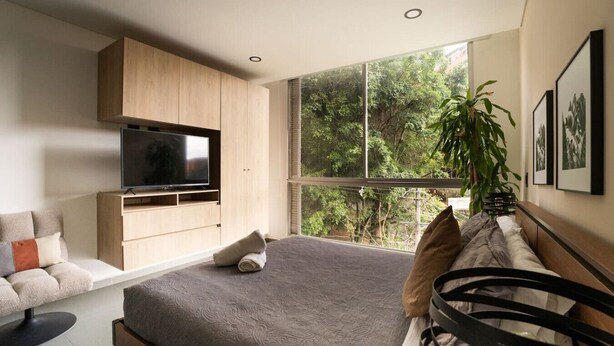 Un dormitorio moderno con una cama grande, un televisor montado en un mueble de madera, un sofá y un ventanal con vistas a árboles.