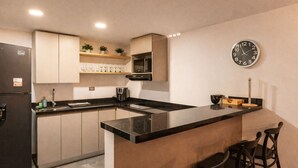 Una cocina compacta con cubierta negra, un refrigerador, un microondas y un reloj en la pared.