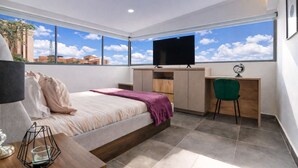 Un dormitorio moderno con cama, escritorio, silla, televisión y ventanas amplias con vista a la ciudad.