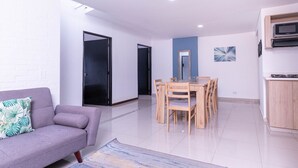 A modern living room with a grey sofa, a wooden dining table with chairs, and a kitchen area with a microwave.