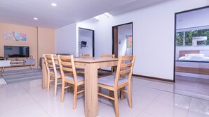 A modern living room with a wooden dining table and chairs, a flat-screen TV, and a large window with a view of greenery.
