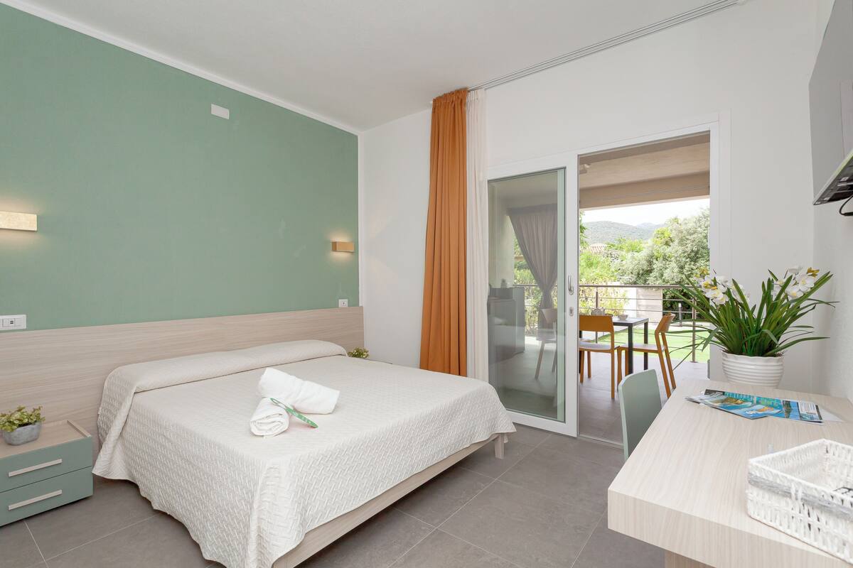 A bedroom with a bed, a desk, a chair, a potted plant, and a sliding glass door leading to an outdoor area.