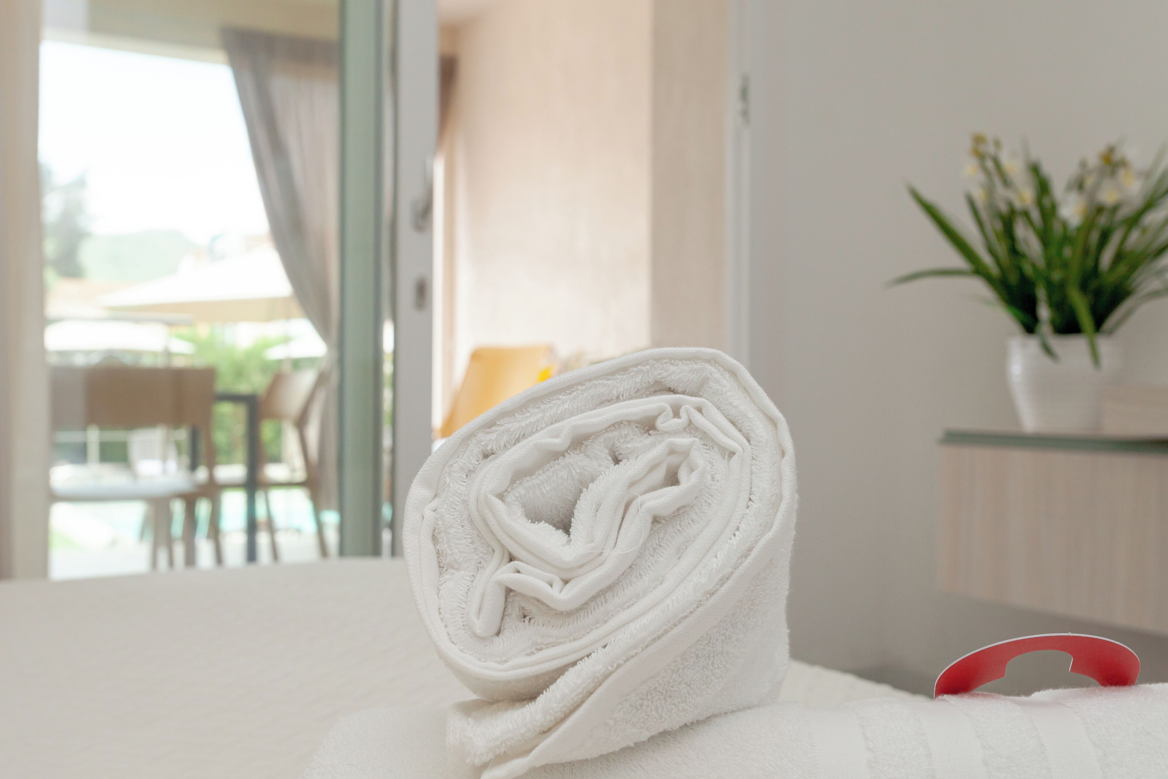 Een netjes opgerolde witte handdoek op een bed, met een rode handdoekhouder, een potplant en uitzicht op een buitenruimte met stoelen en een tafel.