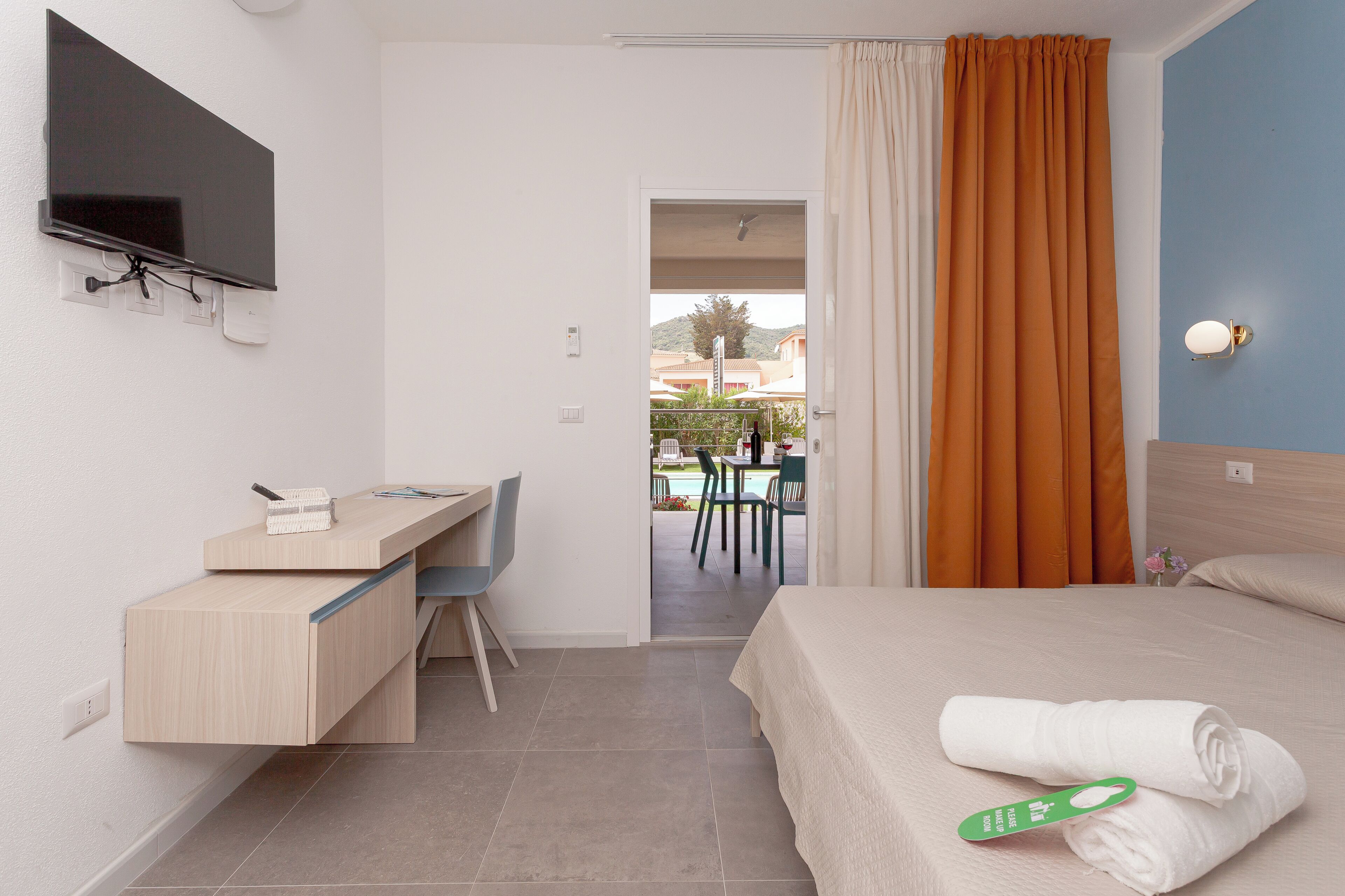 A hotel room with a bed, a desk, a TV, and a view of an outdoor pool area.