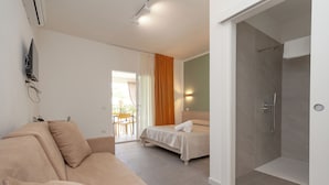 A modern hotel room with a sofa, a bed, a shower area, and a television mounted on the wall.