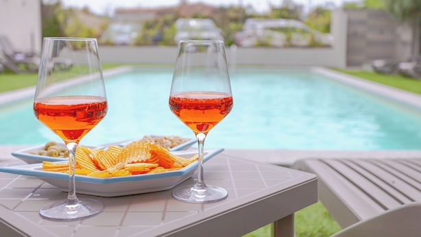 Two glasses of red drink on a table by a pool with a plate of chips.