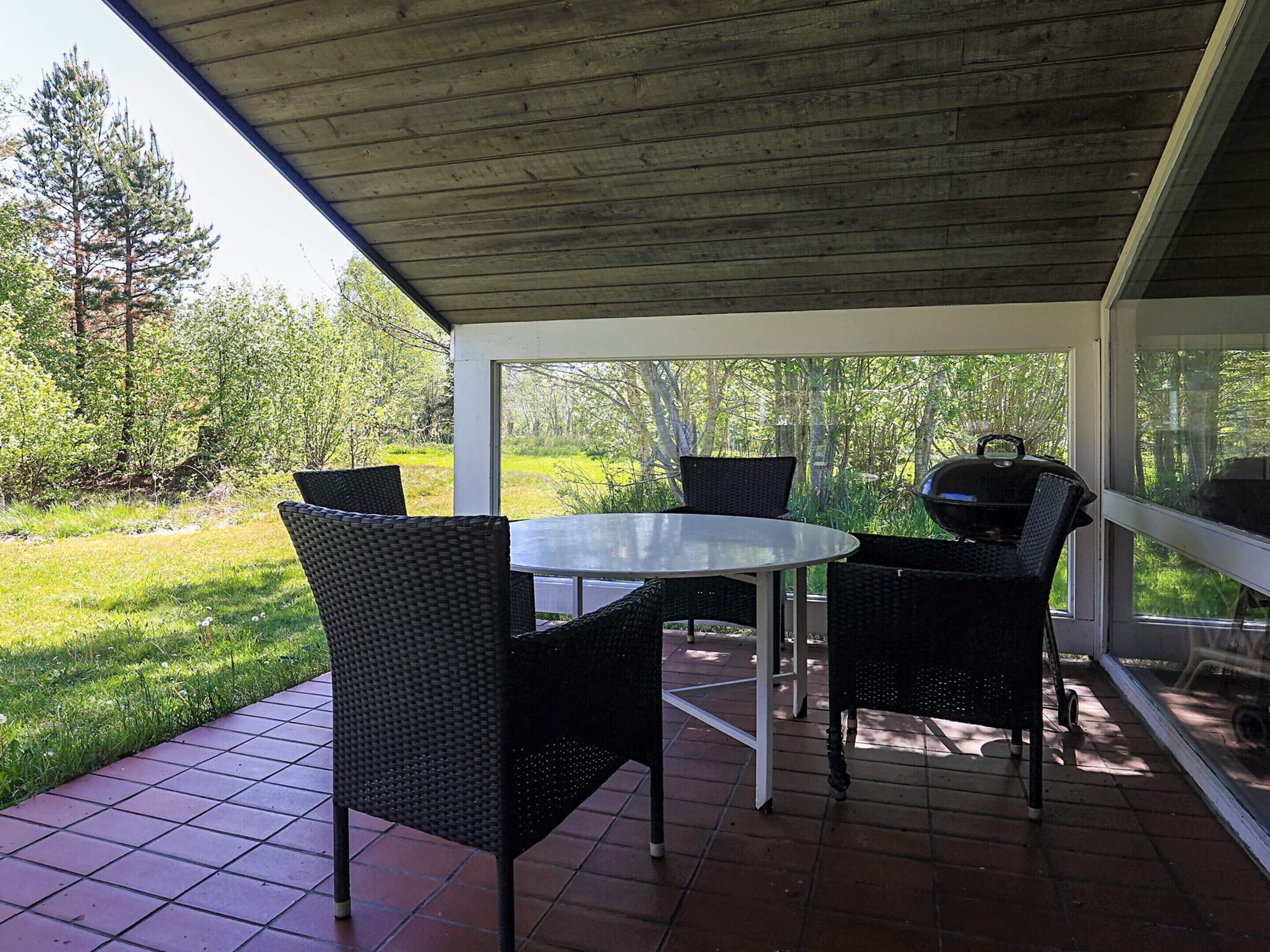 A covered patio with a round table and four chairs, a grill, and a view of a grassy area with trees.