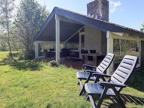 A covered outdoor seating area with a table and chairs, surrounded by grass and trees.