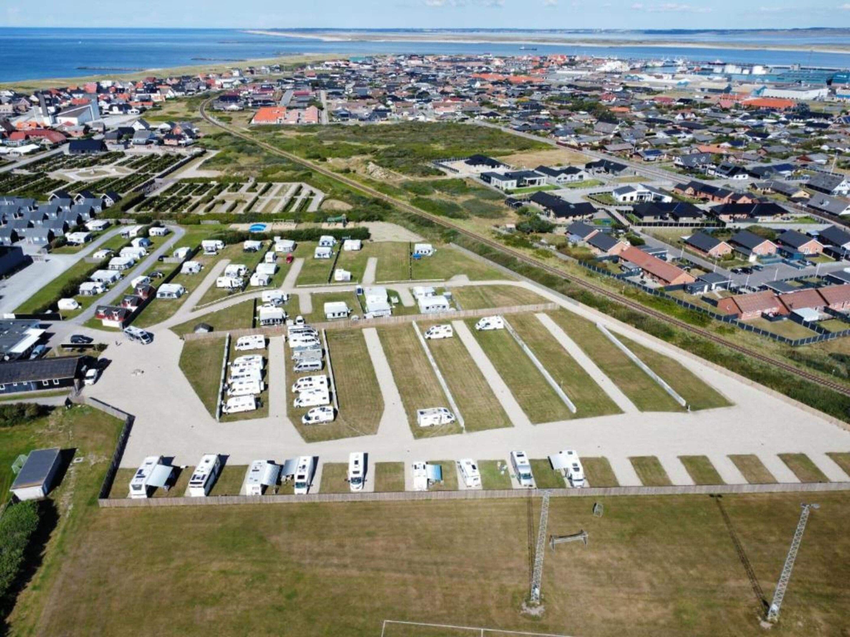 Eine Vogelperspektive eines Wohngebiets mit einem großen Parkplatz und Wohnmobilen, umgeben von Häusern und einem Strand in der Ferne.