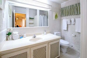 Un bagno con lavandino bianco, uno specchio e un water. Asciugamani appesi al muro e una tenda verde.
