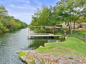 A riverside dock with a covered seating area, surrounded by trees and a grassy bank.