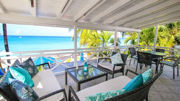 A covered outdoor seating area with wicker furniture and cushions, overlooking a beach and ocean. - Bora Bora Upper 2 Bedroom Apts (Paynes Bay)