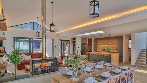 A spacious living area with a dining table set for six, a fireplace, and a kitchen in the background.