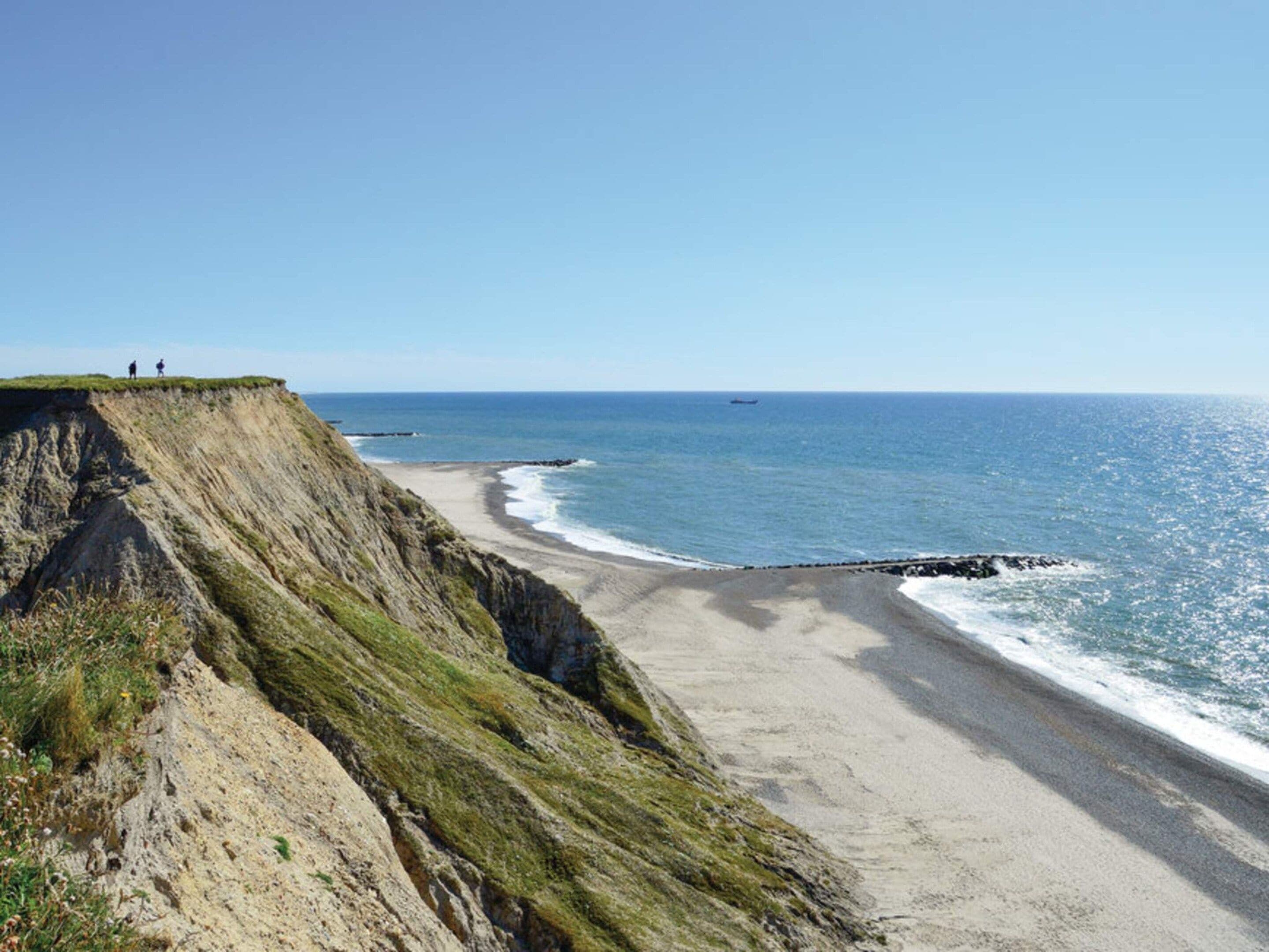 A coastal landscape with a steep cliff, a sandy beach, and the ocean.