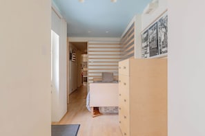 Ein schmaler Flur, der in ein Schlafzimmer mit Bett, Kommode und Wandregal führt.