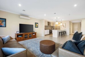Un salon moderne avec un canapé, un pouf et un téléviseur à écran plat. On aperçoit, au fond, une cuisine dotée d’un îlot central et de tabourets de bar.