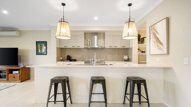 Una cocina moderna con una isla central, tres taburetes de bar, iluminación colgante y un televisor de pantalla plana.