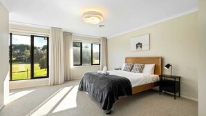 Un dormitorio con un ventanal grande, una cama con cabecera de madera, una mesita de noche con lámpara y un cuadro enmarcado en la pared.