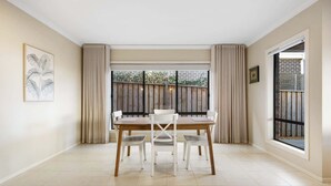 Un comedor con una mesa de madera y sillas blancas, ventanas amplias con cortinas, e impresión enmarcadas con motivos botánicos en la pared.
